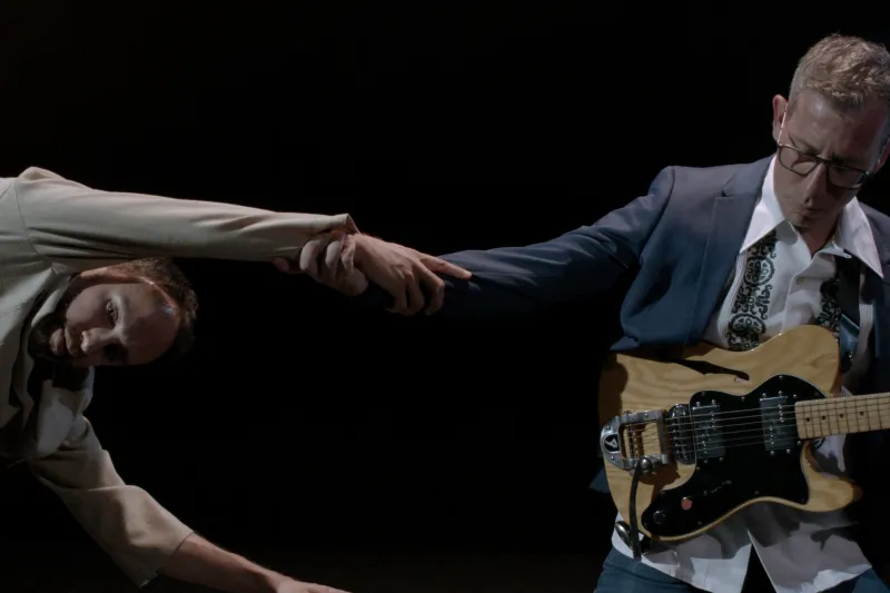 Photo de deux comédiens, l'un avec une guitare, l'autre en mouvement, qui se tiennent par la main